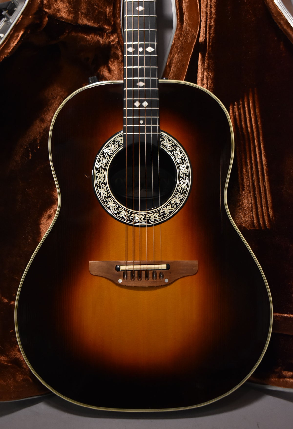 Full front view of a sunburst acoustic guitar with a decorative soundhole rosette and diamond fretboard inlays, resting in a brown velvet case.