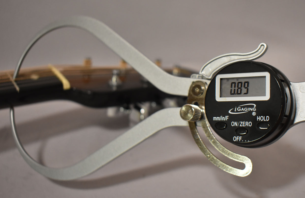 Digital calipers measure the guitar's nut, showing a reading of 0.89, with the headstock and strings blurred in the background.
