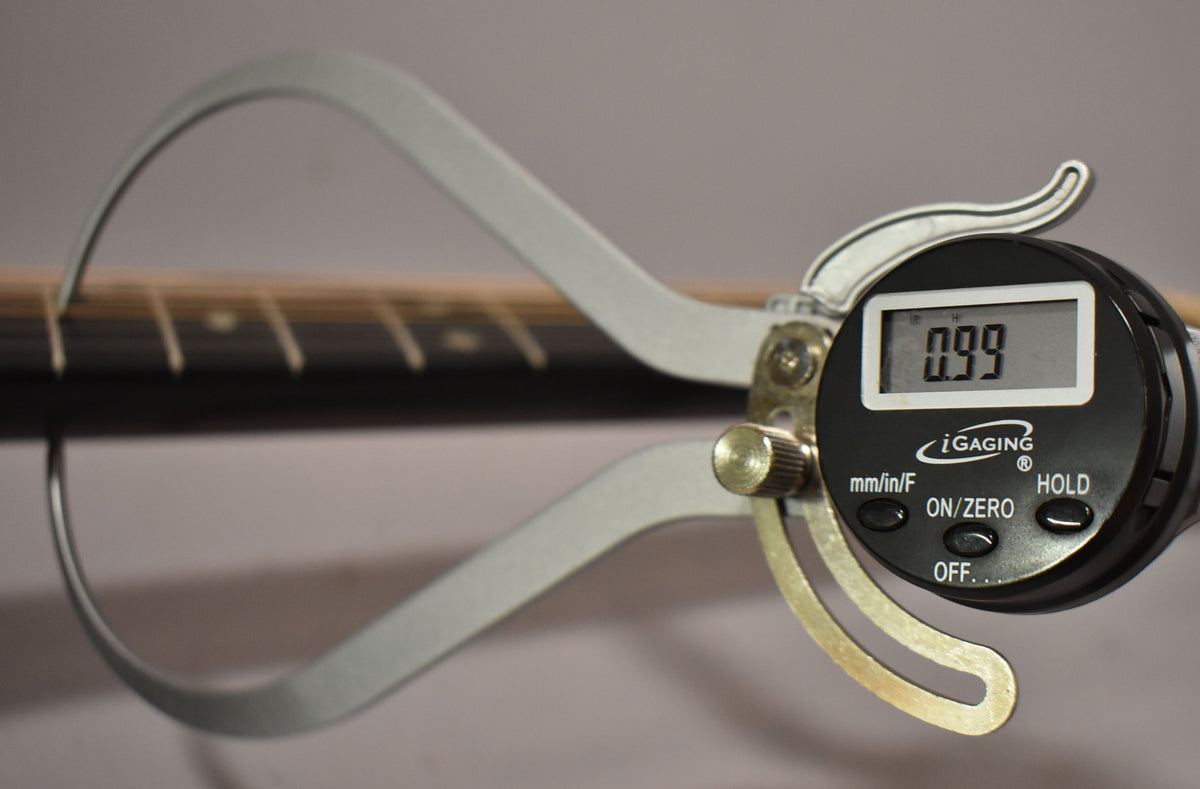 A digital caliper measures the guitar's fret height at 0.99mm, showing the dark fretboard and light frets.