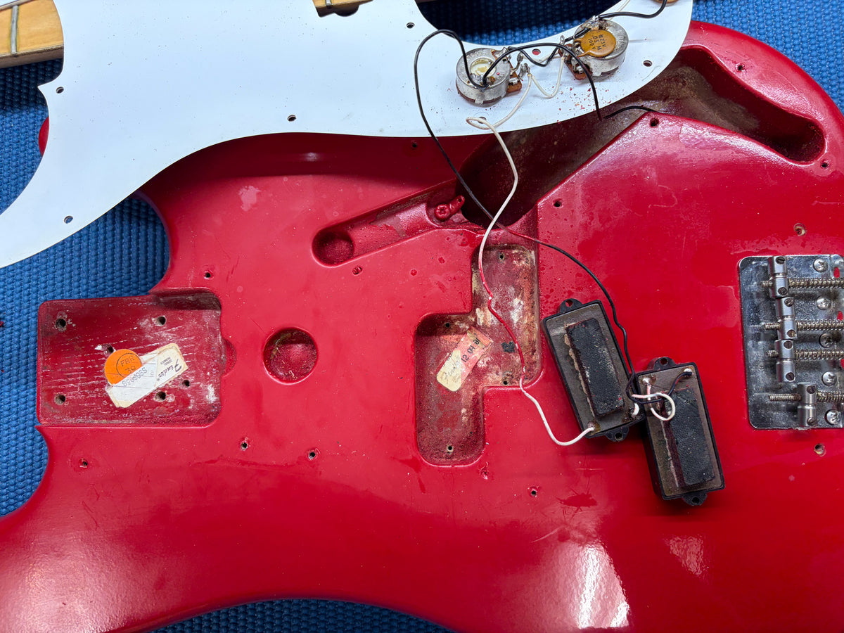 Overhead view of the red bass body with the white pickguard removed, showing the pickups, wiring, and bridge.
