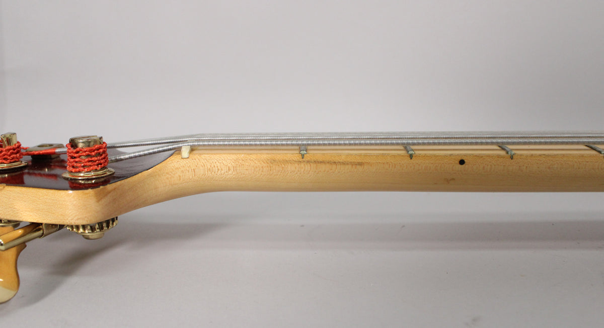 The bass neck, angled from the headstock, shows a light wood finish, dark fretboard, and gold tuners with red string wraps.