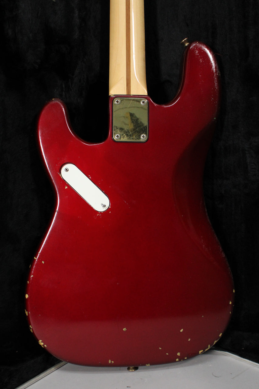 The back of a candy apple red bass shows a maple neck, a worn gold neck plate, a white control cover, and many paint chips.