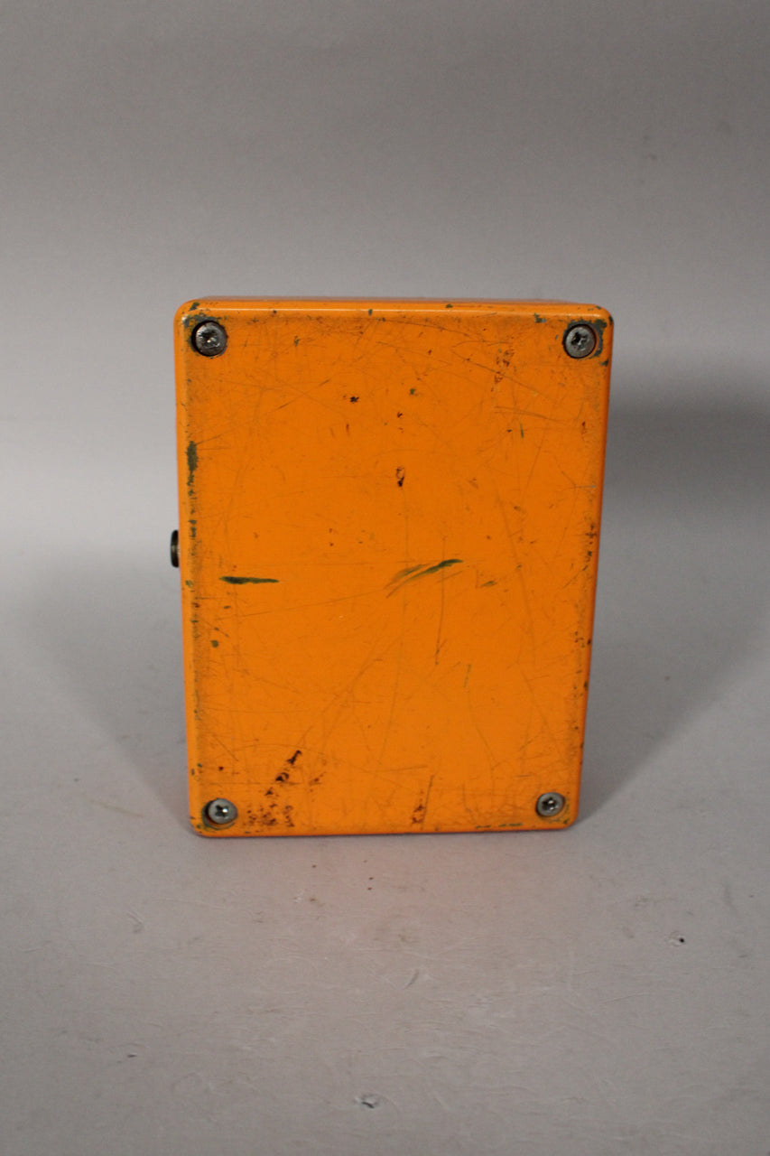 The orange pedal is viewed from the top, showing its scratched surface and four corner screws, with a black knob visible on the left side.