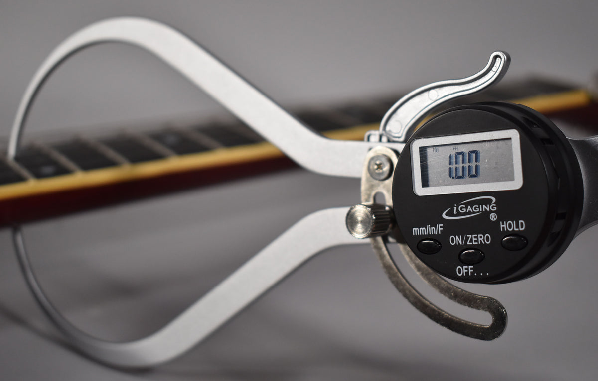 A digital caliper measures the guitar's cherry-finished neck, showing "1.00" on its screen, with fretboard and frets visible.