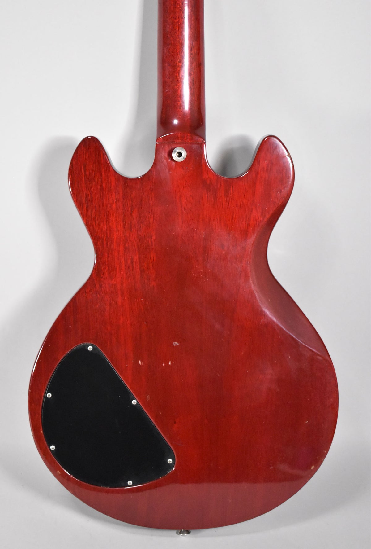 The back of a cherry red electric guitar, showing the neck joint, a black control cavity cover, and minor surface wear.