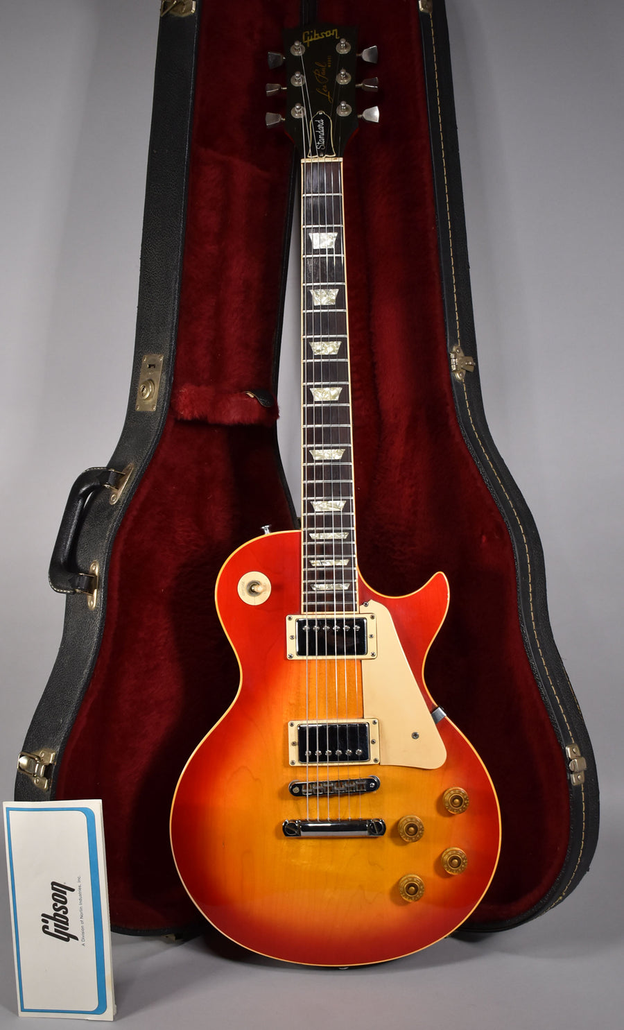 Full-length view of a cherry sunburst guitar with cream binding, gold hardware, and a black headstock, standing in its open, red-lined case.