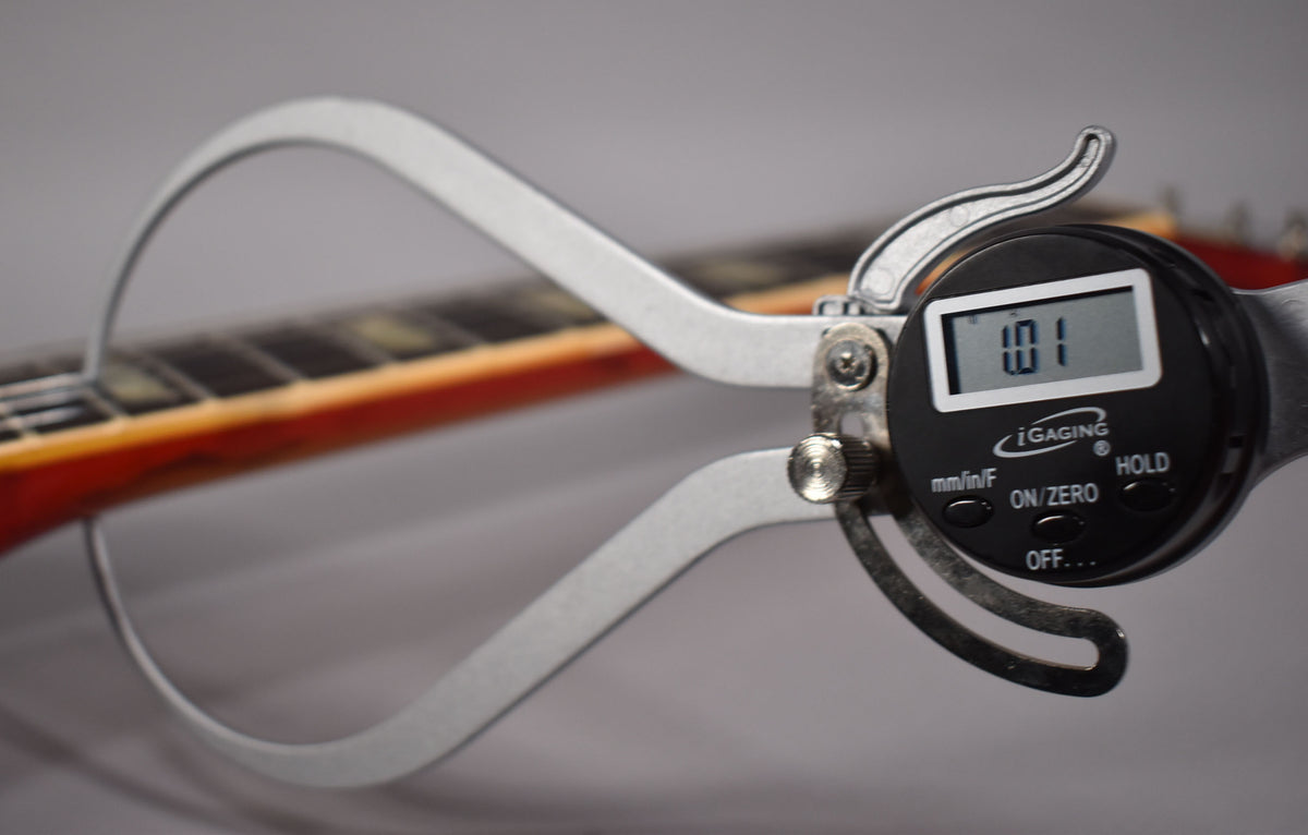 A digital caliper measures the neck thickness of the cherry sunburst guitar, showing "1.01" on its display.