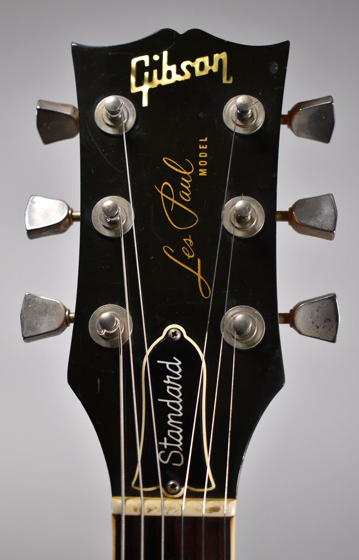 Close-up of a black headstock with gold lettering, six tarnished chrome tuners, and light scratches across the surface.