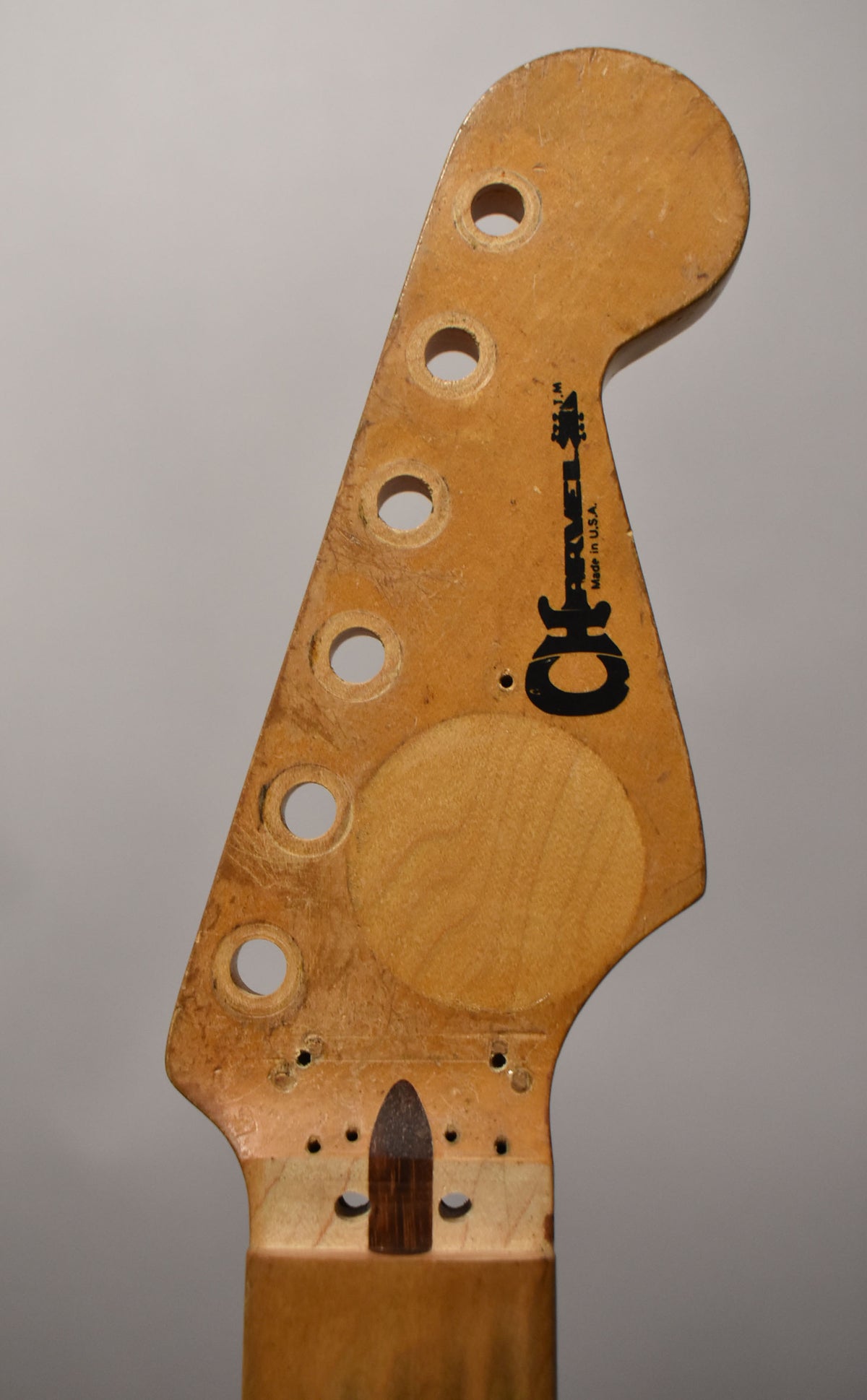 Close-up of a light maple Strat-style headstock with six tuner holes, a circular string tree imprint, and a dark wood bullet truss rod.