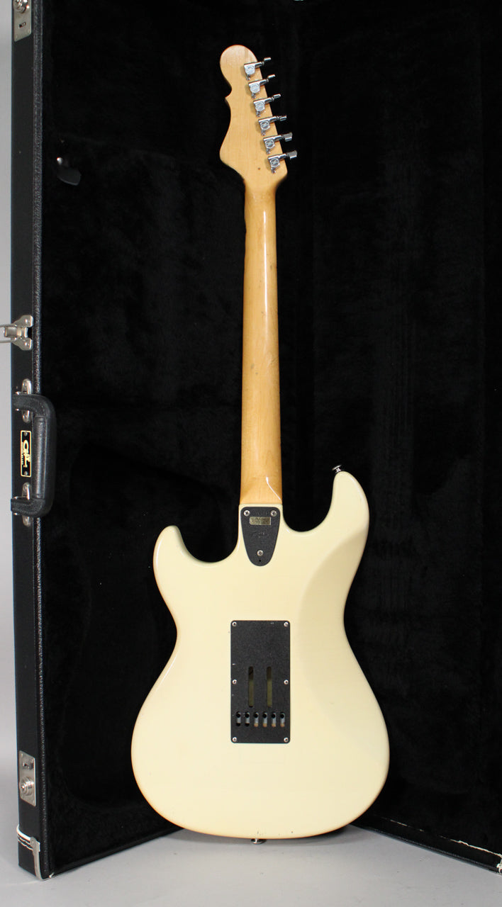 The back of an Olympic White guitar with a maple neck and headstock, showing a black tremolo cover and some finish wear.
