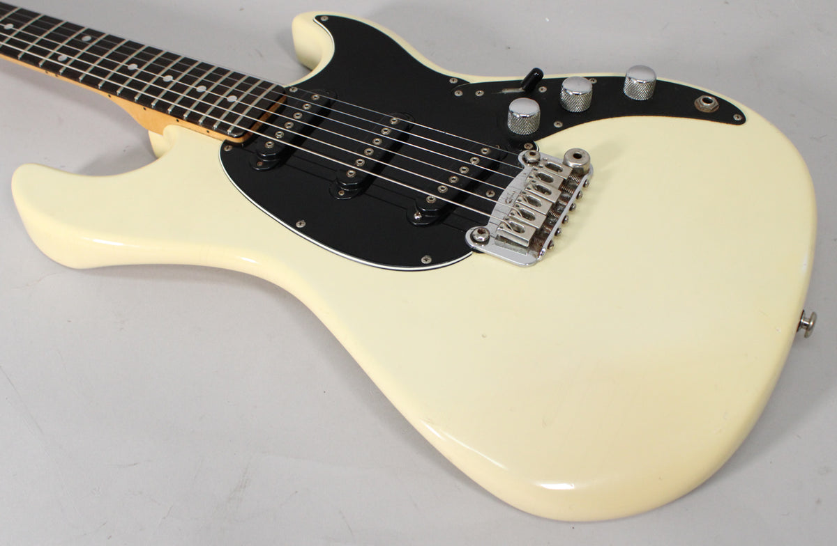 Olympic White electric guitar, angled from above, showing the body, black pickguard, three pickups, chrome bridge, and three control knobs.