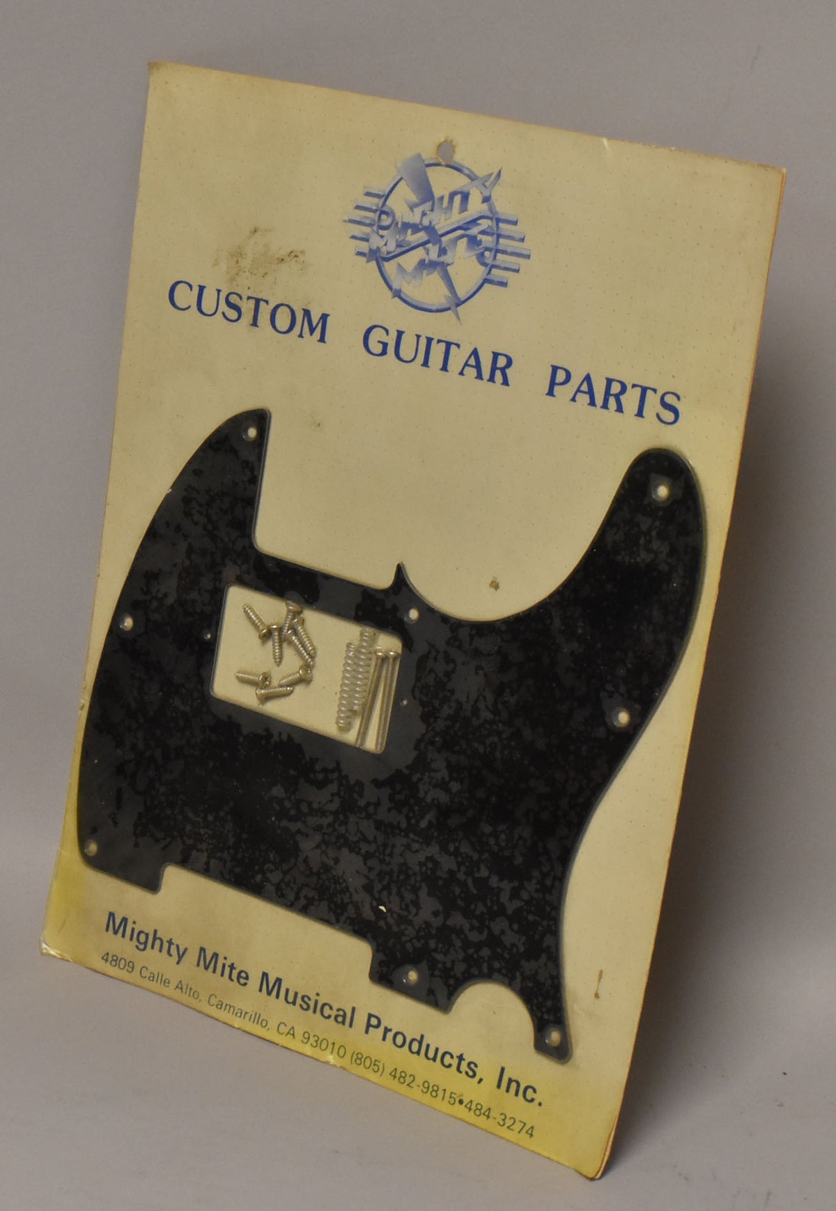 A black T-style pickguard with a subtle mottled texture, still sealed in its original packaging with mounting screws and springs visible.
