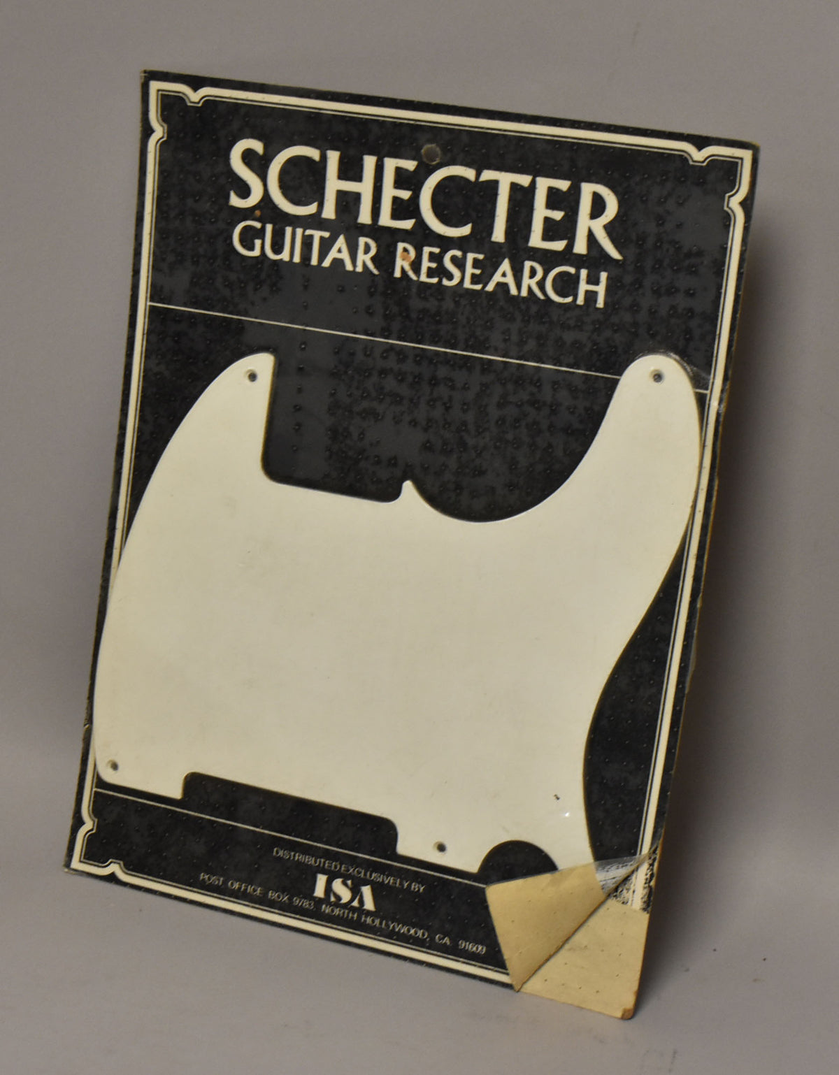 A white pickguard with six screw holes is sealed in clear plastic on a black and white cardboard backing, with a peeled corner.
