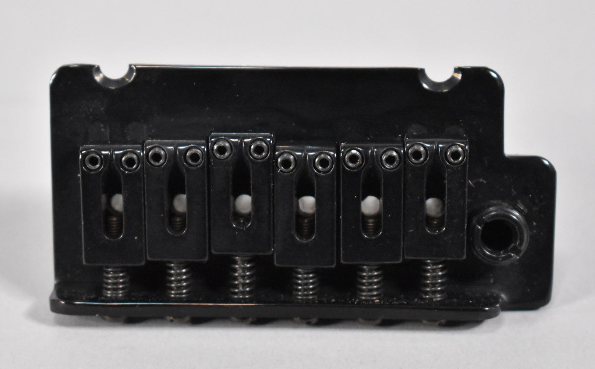 Top-down view of a black tremolo bridge with six saddles, each with a white string guide and adjustment screws.