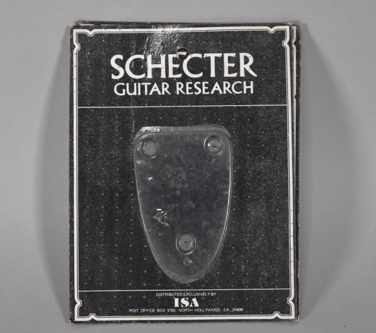 A black, shield-shaped neck plate with three screw holes is visible through clear blister packaging on a dark gray card.