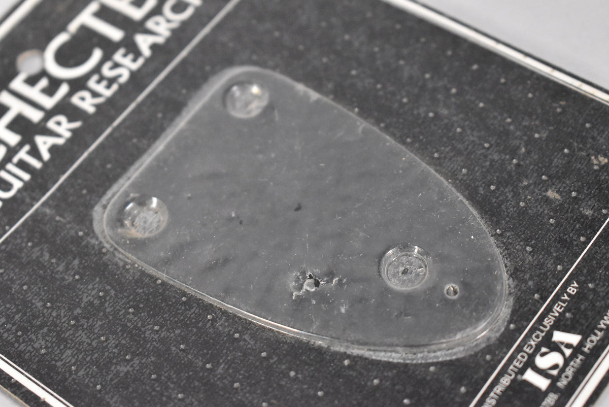 A black, teardrop-shaped neck plate is sealed in clear plastic packaging, with three visible screw holes and a small tear in the plastic.