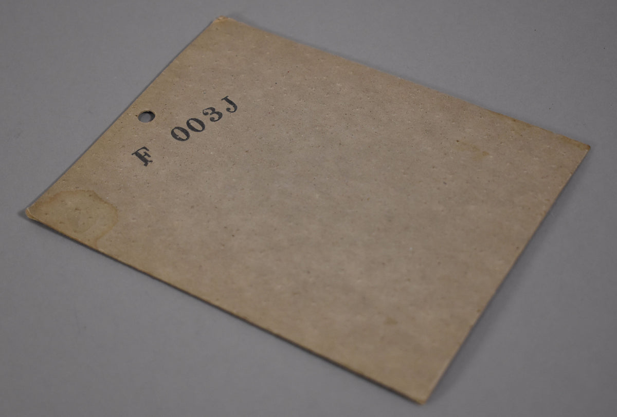 The reverse of the black neck plate is shown, sealed in its original, slightly discolored brown paper packaging with "F 003J" stamped on it.