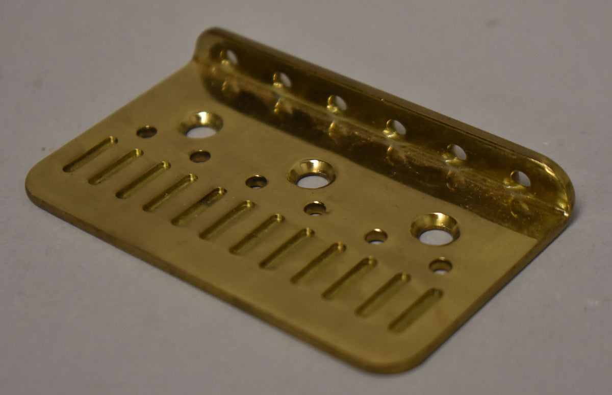 A gold hardtail bridge plate, viewed from above and slightly to the right, showing six string slots and various mounting holes.