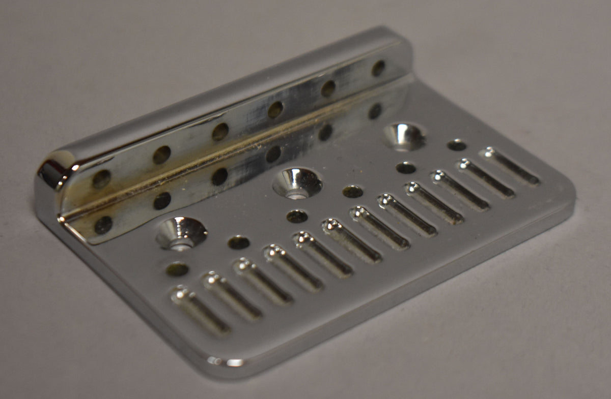 A chrome hardtail bridge plate, viewed from above at an angle, shows six string-through-body holes and some tarnishing on the back edge.