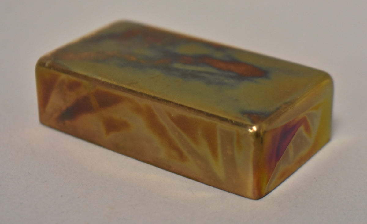 A rectangular gold humbucker cover, angled to show the top and one long side, with iridescent purple and green tarnishing.