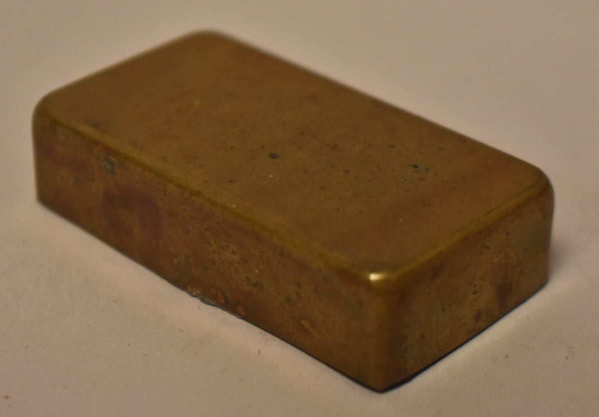 A single gold brass humbucker cover, viewed from above at an angle, showing a worn, tarnished finish with some pitting.