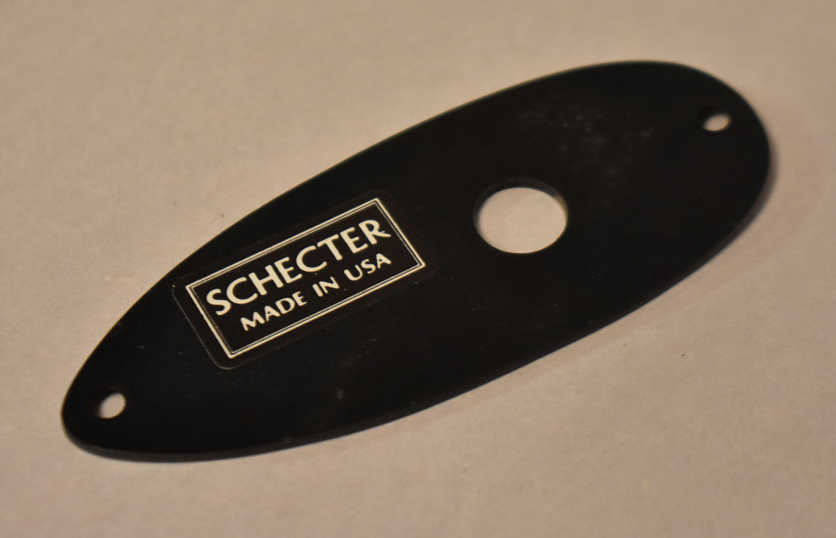 Black oval input jack plate with a central jack hole, two screw holes, and a rectangular "Schecter Made in USA" label.