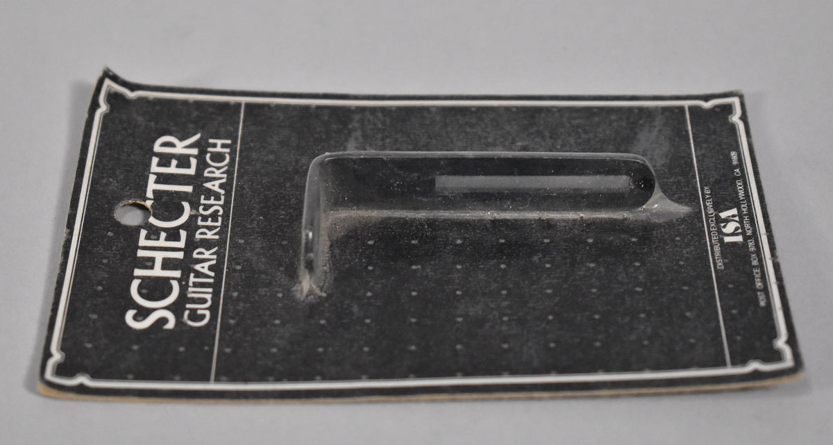A black, rectangular pickguard bracket is sealed in clear plastic on a black and white cardboard backing, viewed from a slight angle.