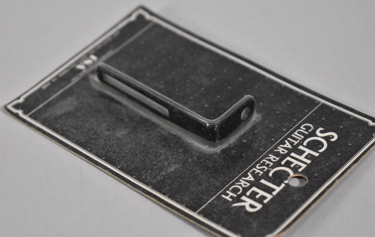 A black L-shaped pickguard bracket with a screw hole is sealed in clear plastic on a dark, textured card.