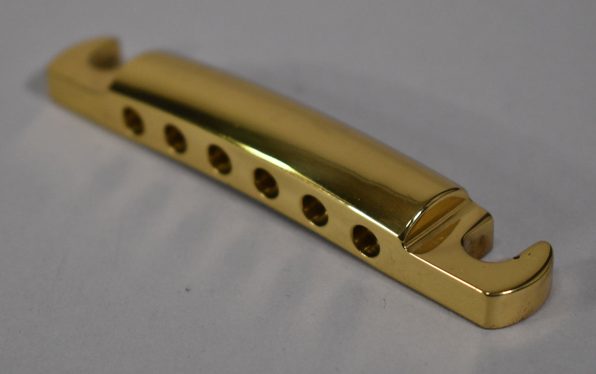 A shiny gold brass stoptail tailpiece, viewed from above and slightly to the side, showing six string holes and mounting hooks.