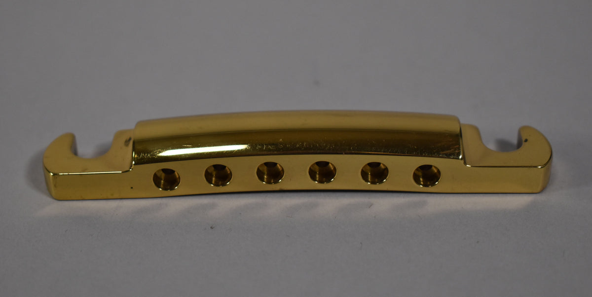 Top-down view of a shiny gold brass stoptail, showing six string holes along the front edge and two mounting hooks.