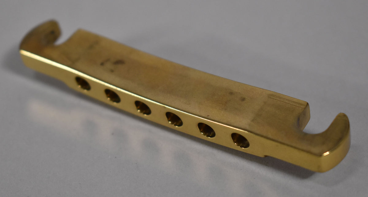 A brass stoptail tailpiece, angled from the left, shows six string holes and a matte finish with some light scratches.