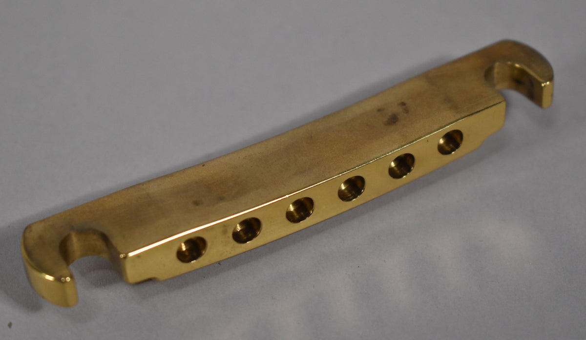 A polished brass stoptail tailpiece, viewed from above, shows six string holes and two mounting hooks.