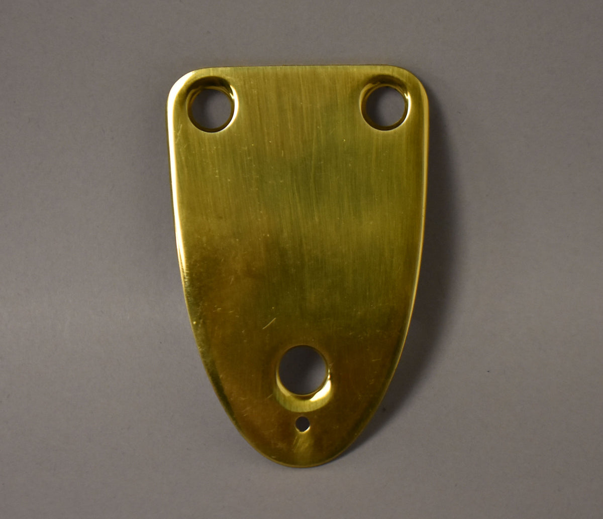 A brass neck plate, viewed from above, has a brushed finish with two holes at the top and one larger hole with a smaller one below it at the bottom.