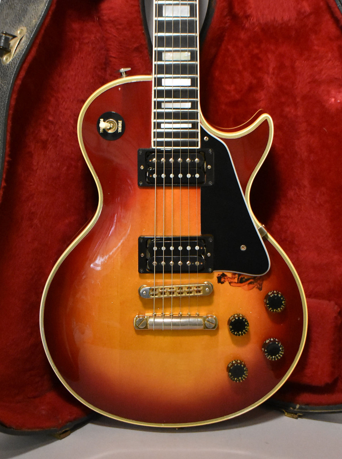 Full body shot of a cherry sunburst Les Paul Custom, showing gold hardware, black pickups, and a black pickguard with a pin-up sticker.