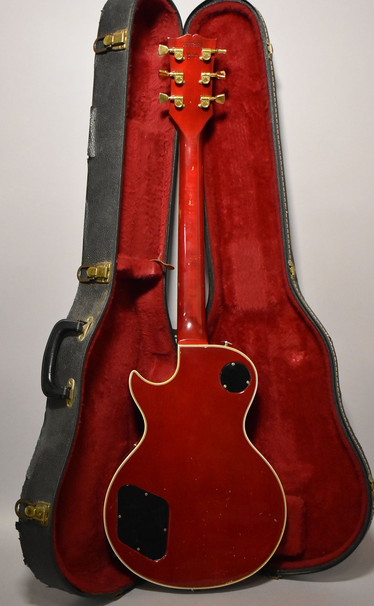 Rear view of a cherry sunburst guitar with gold tuners and strap button, showing light wear and a black control cavity cover, resting in a red-lined case.