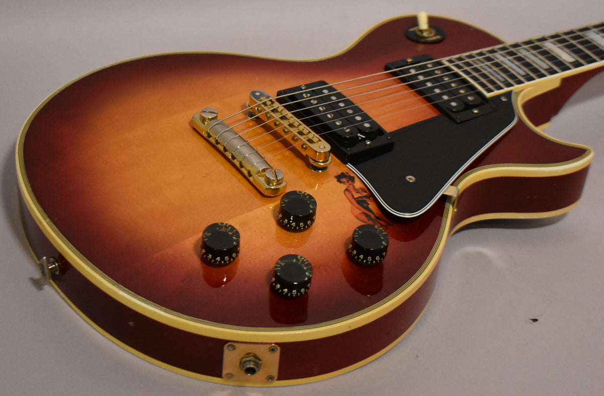 The cherry sunburst guitar body is angled, showing the gold bridge, black pickups, four black control knobs, and a pin-up girl decal.
