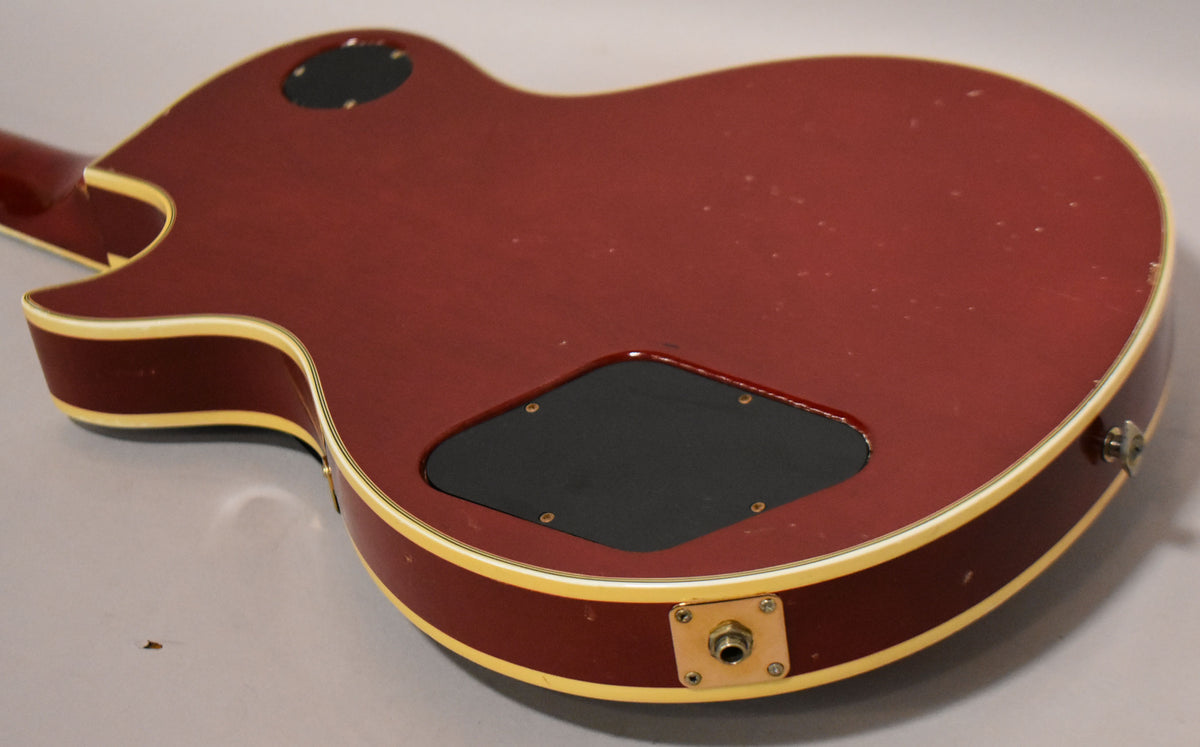 The back of a cherry sunburst electric guitar, showing a black control cavity cover, output jack, and light finish wear.