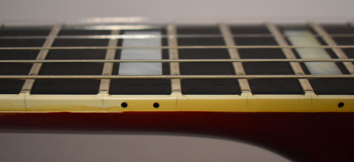 Close-up of the fretboard's lower edge, showing cream binding with visible checking, wear, and dark side dots.