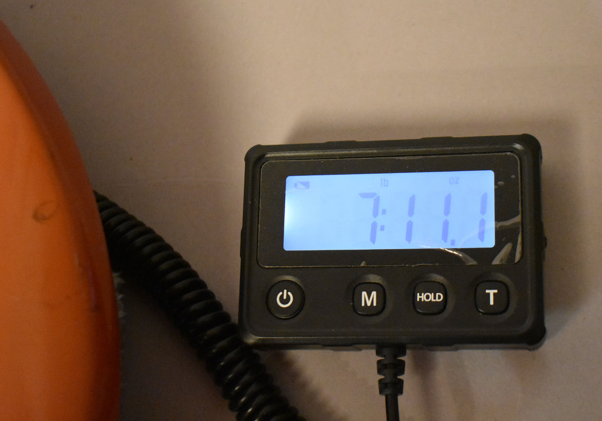 A close-up of a digital scale's screen showing "7:11" in blue, indicating 7 pounds, 11 ounces, with an orange guitar body partially visible left.
