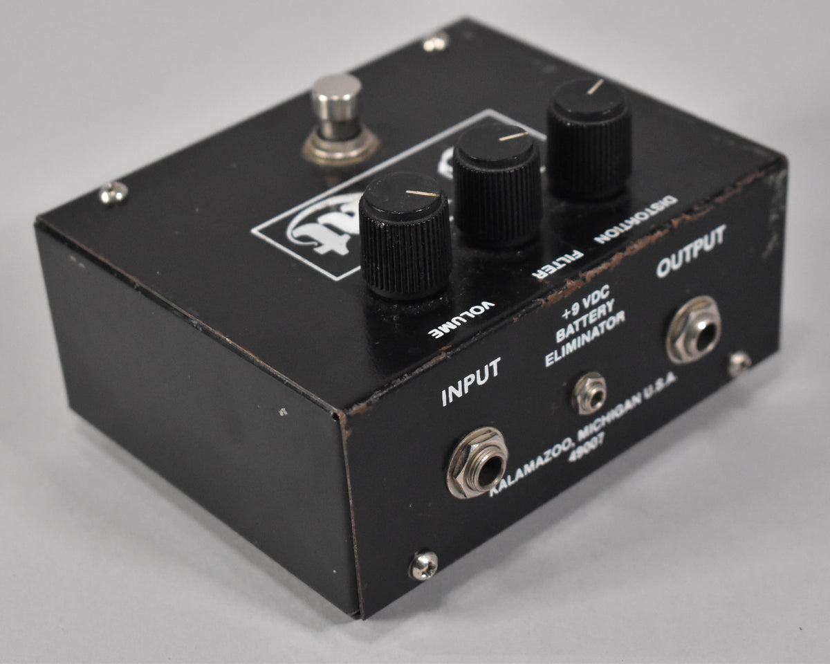 A black metal distortion pedal, viewed from the front right, showing three black knurled knobs, input/output jacks, and edge wear.