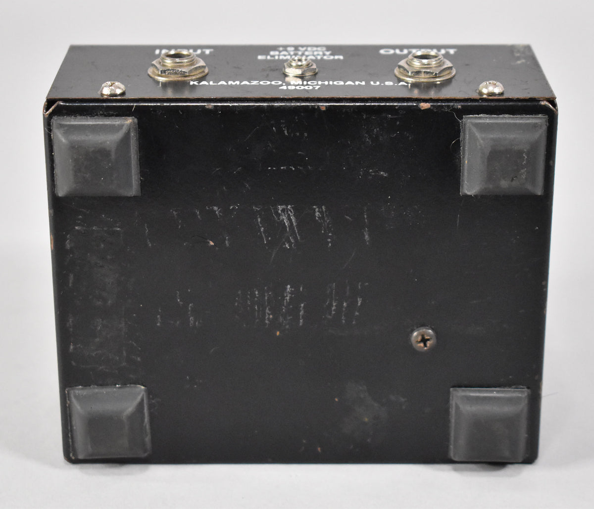 Bottom view of the black pedal, showing four rubber feet, a single screw, and numerous scratches and scuffs on the finish.