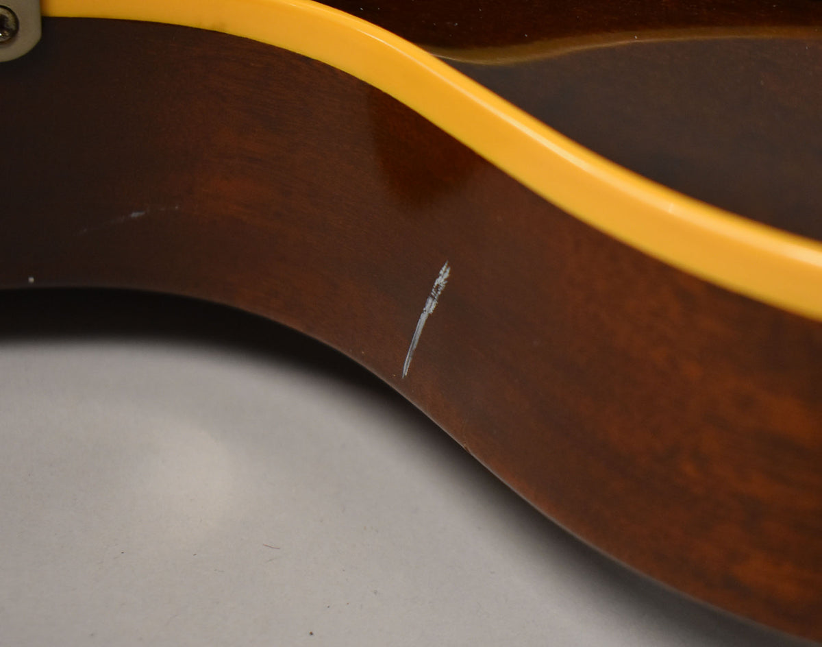 Close-up of a guitar's dark brown body with a yellow binding, showing a prominent white scratch and several smaller marks.