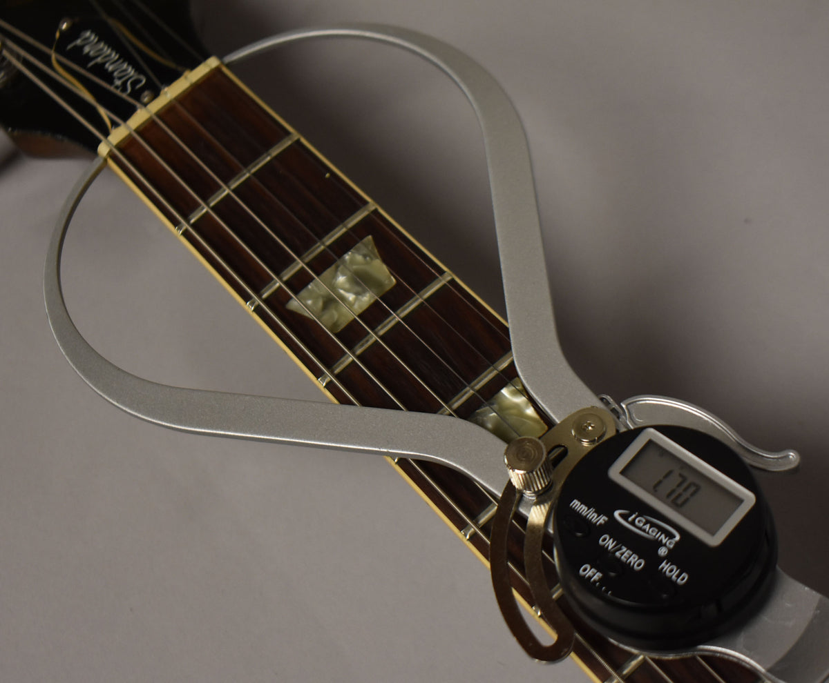 Close-up of the guitar's dark fretboard with trapezoid inlays, a digital fret height gauge showing "1.70" clamped on the 7th fret.