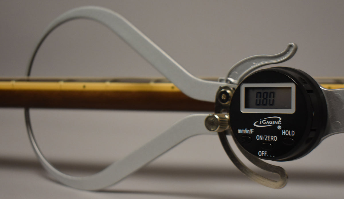 A digital caliper measures the guitar's neck, showing the light brown fretboard with dark dot inlays and a dark brown neck.