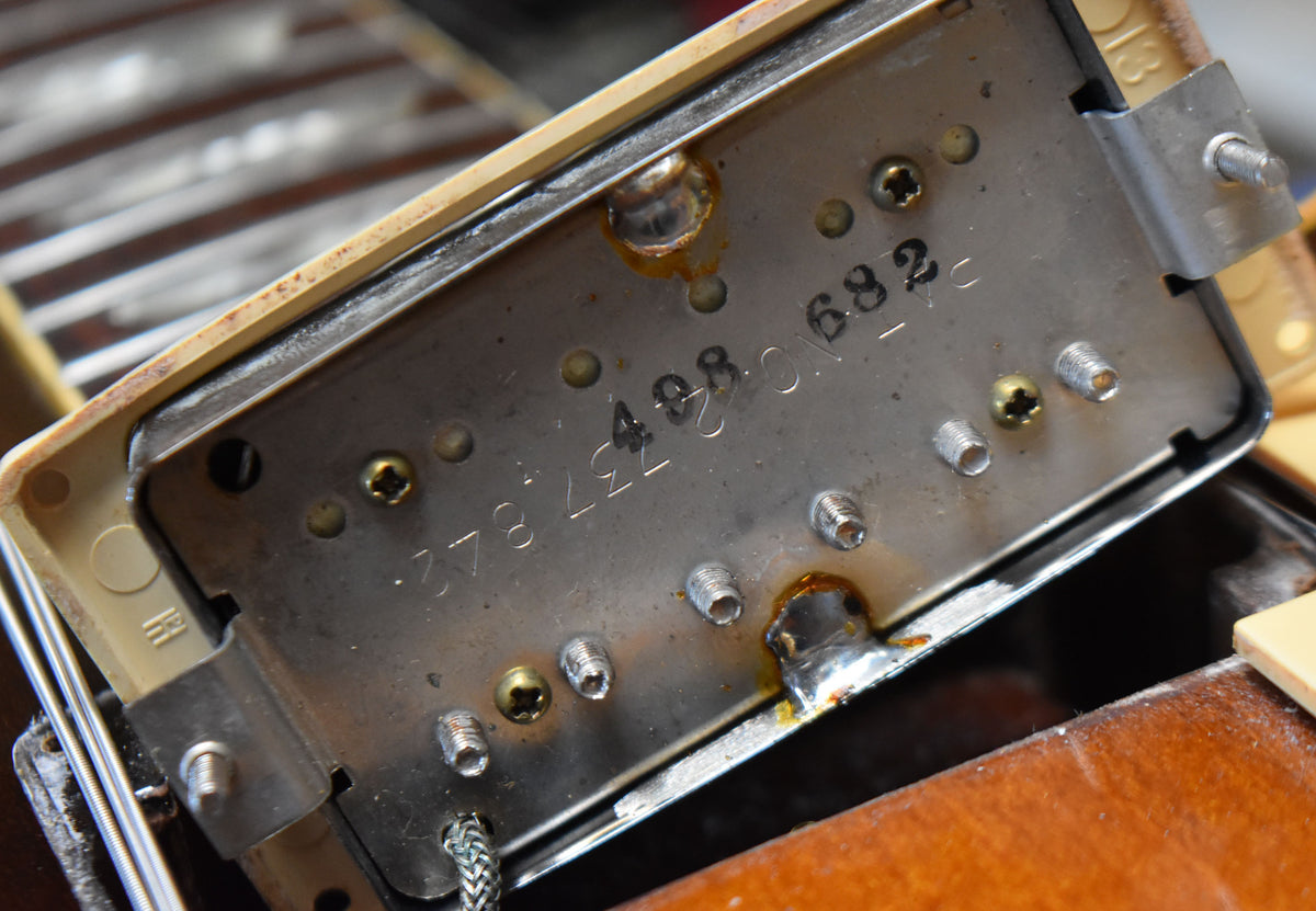 Close-up of the underside of a humbucker pickup, showing the metal baseplate with stamped numbers and solder joints.