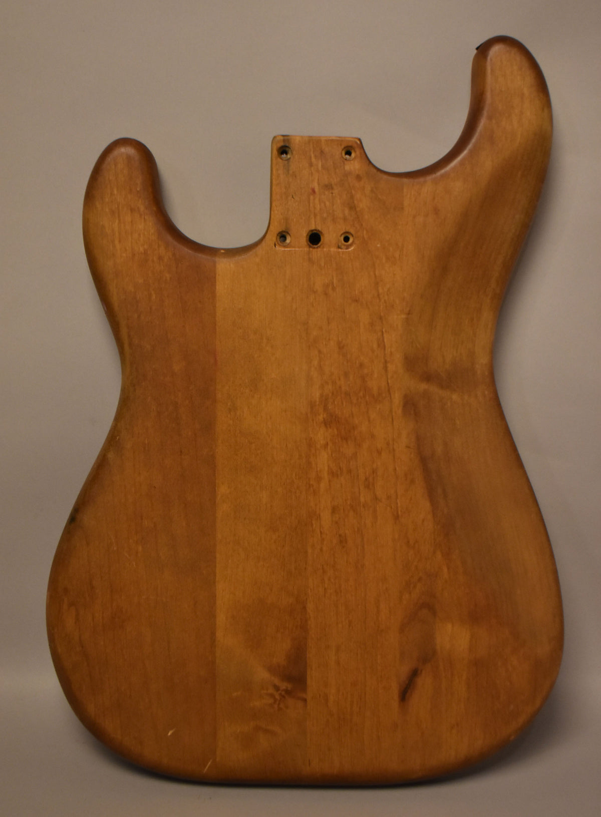 Full rear view of a stripped natural wood Stratocaster body, showing visible grain, some dark spots, and a few light scratches.