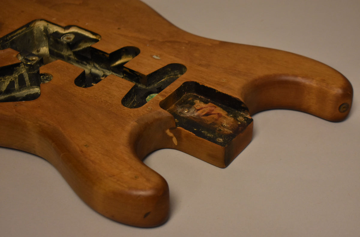 Close-up of a stripped natural Stratocaster body, showing the control cavity with some remaining black paint and a small chip.