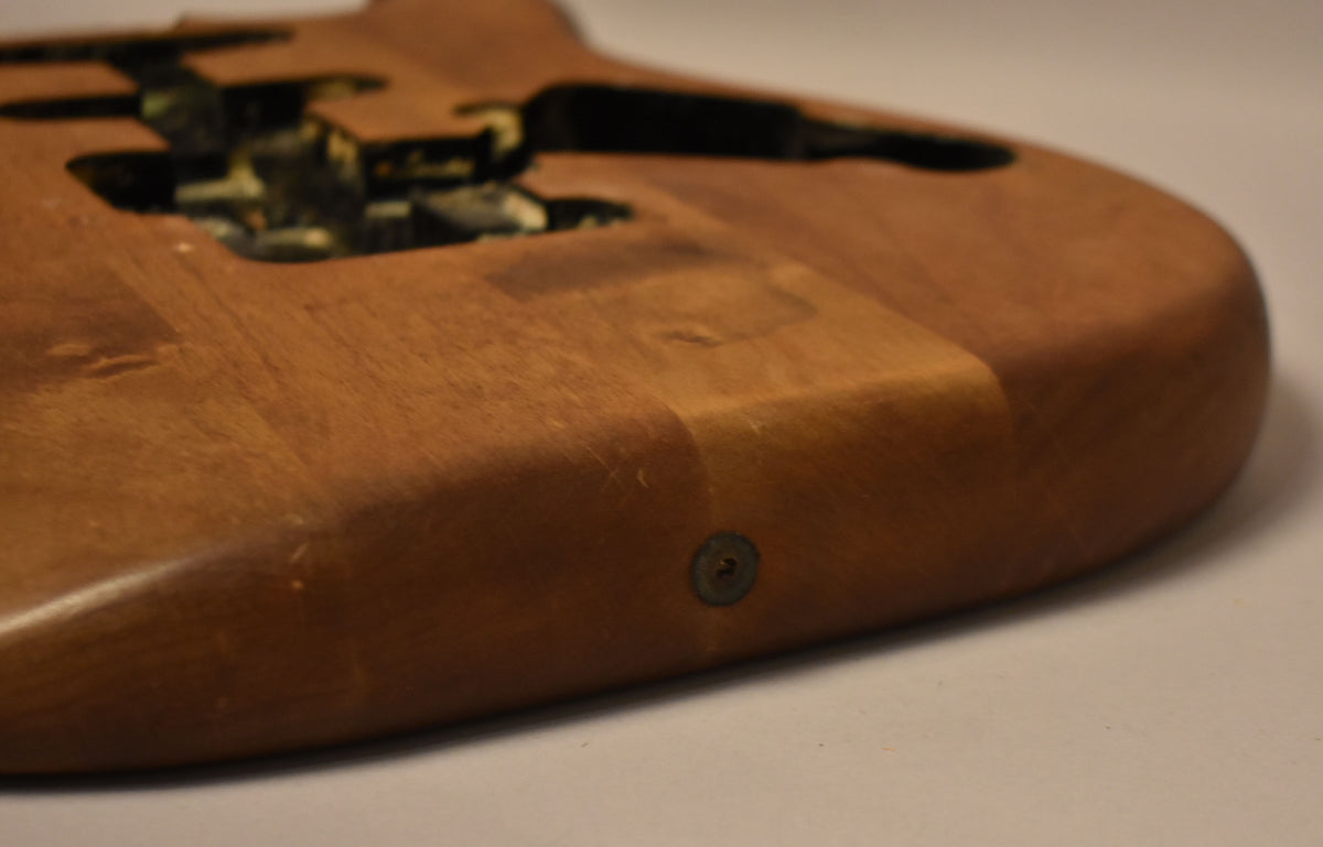 Close-up of the lower bout of a stripped natural Stratocaster body, showing wood grain, light scratches, and a strap button.