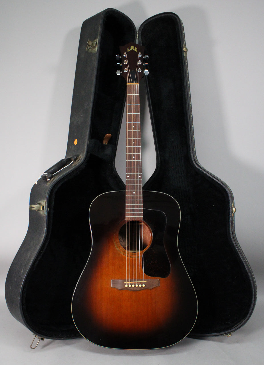 Full-length view of a sunburst acoustic guitar with a dark headstock and pickguard, standing in an open black hard case.