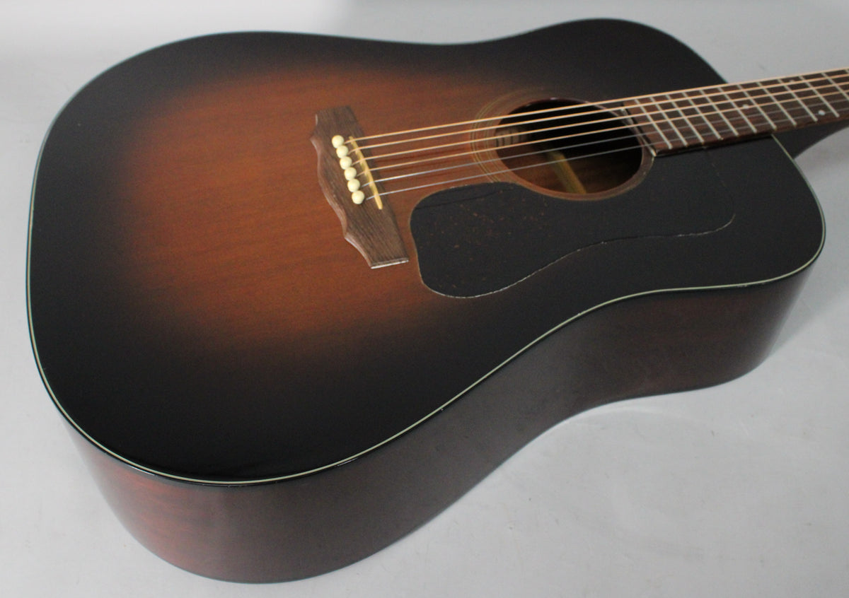 The sunburst acoustic guitar is angled from the upper left, showing the dark brown body, pickguard, bridge, and six strings.
